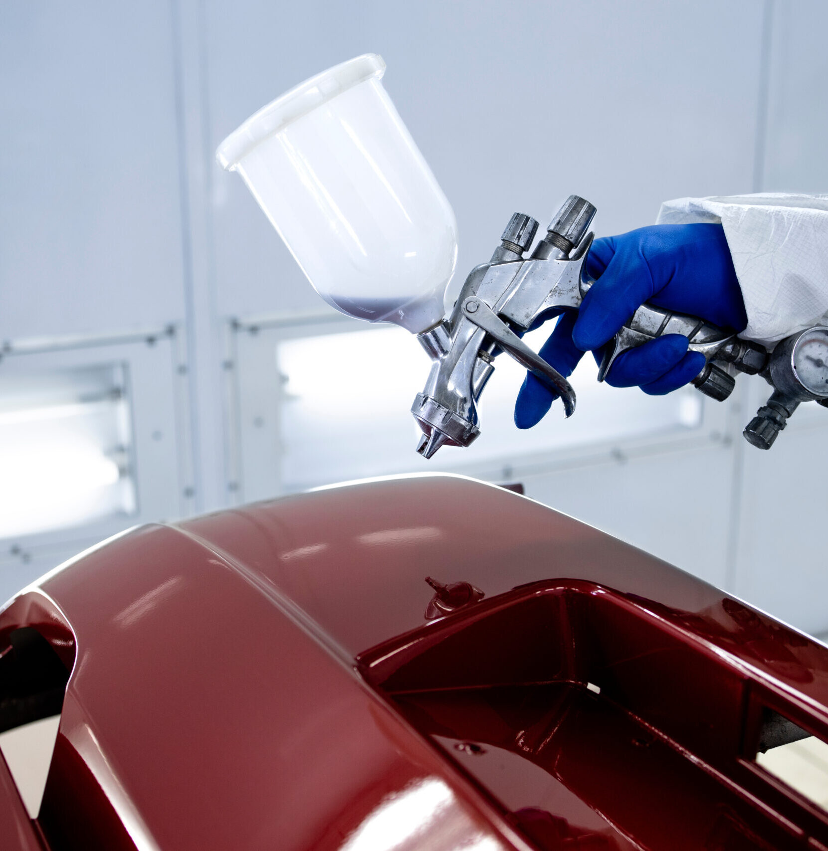 Car painter in protective clothes and mask painting automobile bumper with metallic paint and varnish in chamber workshop.
